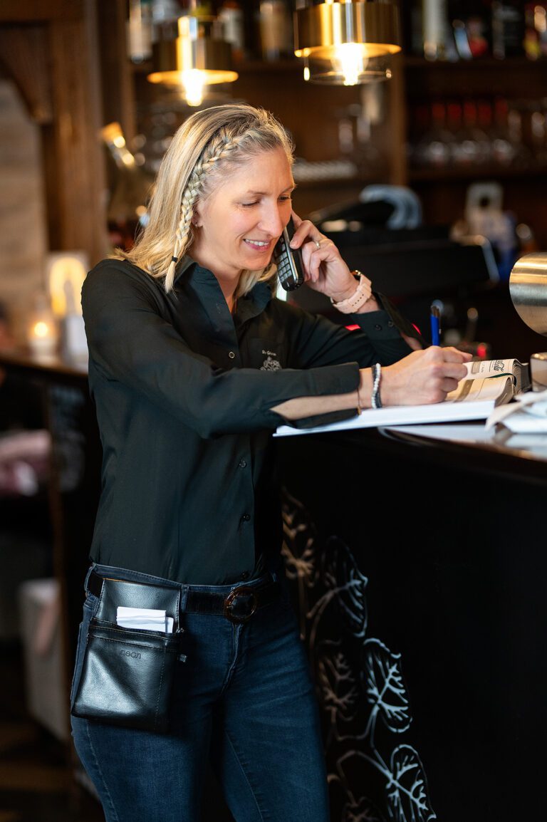 Mitarbeiterin im Restaurant mit Funkgerät am Ohr, lächelnd beim Telefonat und Notizen aufnehmend am Tresen.