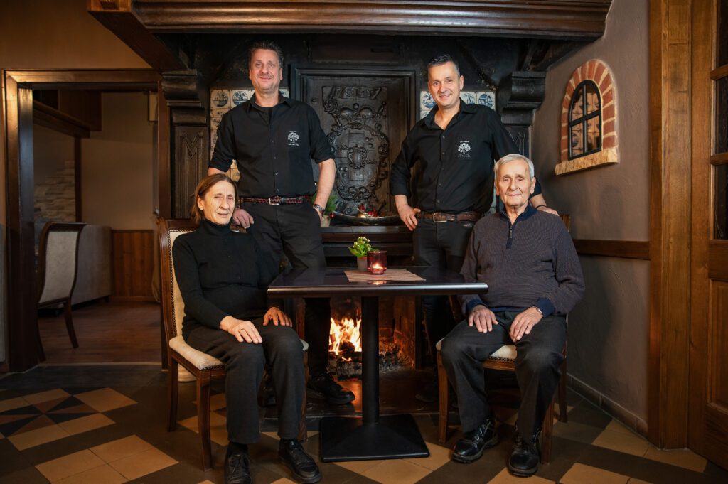 Mehrgenerationenfoto im Restaurant „Der Grieche unter den Linden“ – Gründerfamilie und Nachfolger vor einem offenen Kamin.
